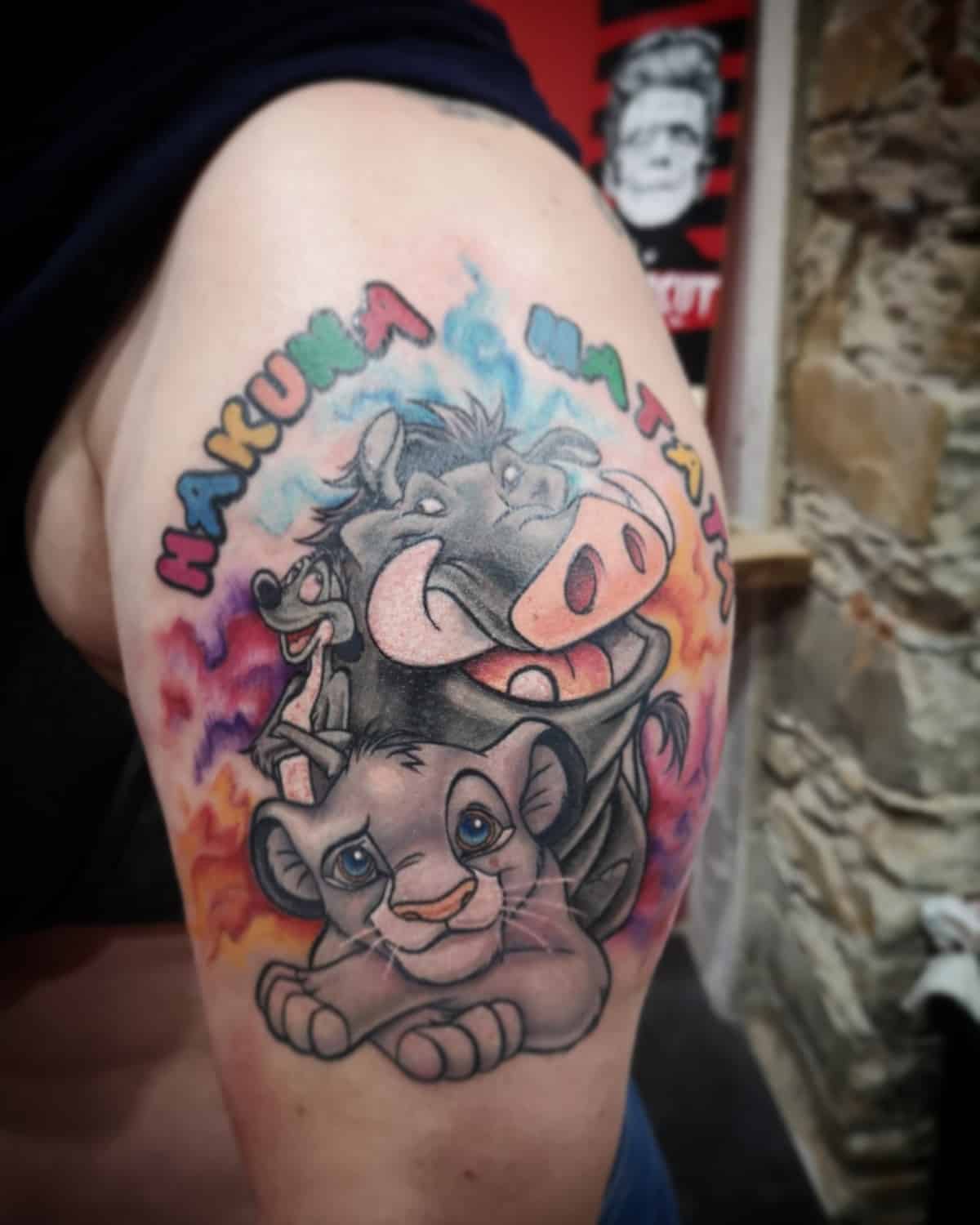 Colorful tattoo of Simba, Timon, and Pumbaa with "Hakuna Matata" on an upper arm